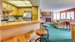 Cozinha compacta com ilha central, banquetas e sala de estar com lareira.