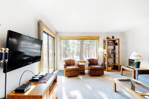 A living room with a TV, leather chairs, a bookshelf, and a view of trees. - Condo with Shared Hot Tubs, Pool, and Sauna & Great Mountain Views (Mammoth Lakes)