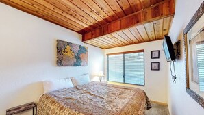 Een slaapkamer met een houten plafond, een bed met een beddeken met patroon, een telefoon aan de muur en een raam met zonwering.