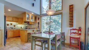 A spacious kitchen with wooden cabinets, a dining area with a table and chairs, and a large window providing a view of trees. - Two-level, forest-view condo with deck - steps from the Village & Ski Back Trail (Mammoth Lakes)