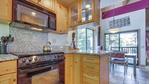 A kitchen with wooden cabinets, a stove, granite countertops, and a dining area with a chair and table. - Two-level, forest-view condo with deck - steps from the Village & Ski Back Trail (Mammoth Lakes)