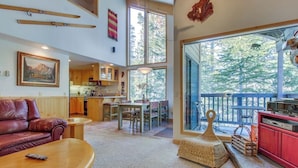 A spacious living area with a large window, a cozy fireplace, and a dining area. - Two-level, forest-view condo with deck - steps from the Village & Ski Back Trail (Mammoth Lakes)