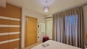 Quarto com cama, guarda-roupa, espelho, um banquinho vermelho e janela com cortinas.
