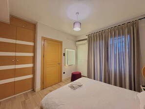 Ein Schlafzimmer mit einem Bett, einem Schrank, einem Spiegel, einem roten Hocker und einem Fenster mit Vorhängen.