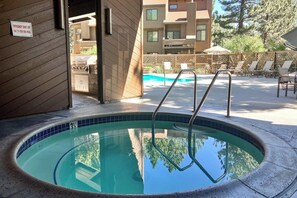 A swimming pool with a metal ladder, a sign indicating an emergency shut-off switch for the spa pool, and a view of a building with a patio area. - Charming Ground-Level Condo with Shared Hot Tub, Pool, Sauna, & Tennis Courts (Mammoth Lakes)