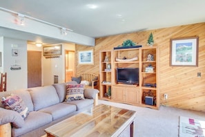 A living room with a sofa, coffee table, wooden entertainment unit, and wall-mounted TV. - Charming Ground-Level Condo with Shared Hot Tub, Pool, Sauna, & Tennis Courts (Mammoth Lakes)
