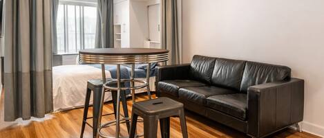 A modern living room with a leather sofa, a round table with stools, and a bed with a white headboard.