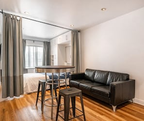 Ein modernes Wohnzimmer mit einem Ledersofa, einem runden Tisch mit Hocker und einem Bett mit weißem Kopfteil.