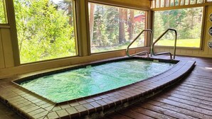 Un jacuzzi con borde de azulejos, cubierta de madera y ventanas amplias con vista al bosque.