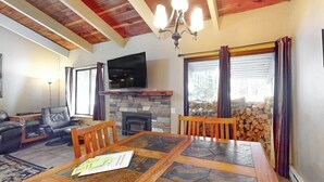 A living room with a stone fireplace, a television, a dining table, and a fireplace mantel. - Modern condo with WiFi, fireplace, pool, & hot tub - near local attractions (Mammoth Lakes)