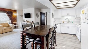 A modern kitchen with white cabinets, a dining area with a wooden table and chairs, and a living room with a sofa and a TV. - Charming mountain condo w/ mountain views, shared pool & hot tub (Mammoth Lakes)