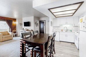 Eine moderne Küche mit weißen Schränken, ein Essbereich mit Holztisch und -stühlen sowie ein Wohnzimmer mit Sofa und Fernseher.