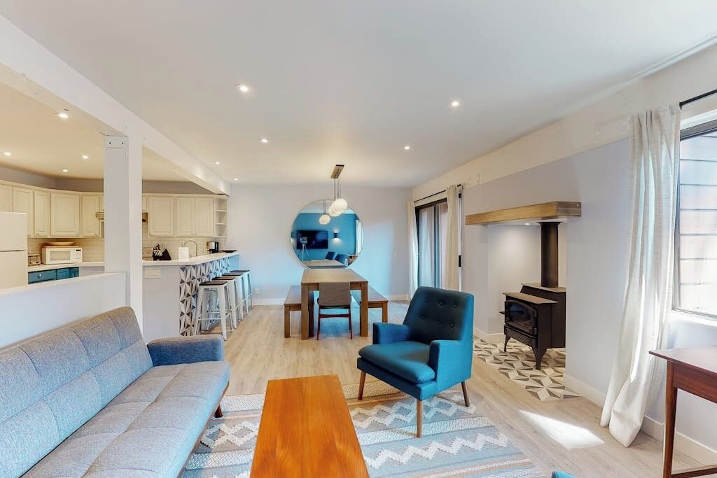 A modern living room with a sofa, a blue armchair, a wooden coffee table, and a kitchen area in the background.