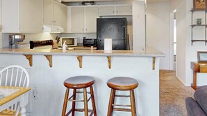 A kitchen with white cabinets, a black refrigerator, a microwave, a stove, and two wooden stools. - Comfy Condo Close to Skiing with Fireplace, Seasonal Pool, Hot Tub, & WiFi (Mammoth Lakes)