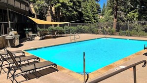 A swimming pool with lounge chairs and a shaded area, surrounded by trees and a fence. - Comfy Condo Close to Skiing with Fireplace, Seasonal Pool, Hot Tub, & WiFi (Mammoth Lakes)