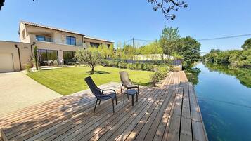Une terrasse en bois avec deux chaises et une table, une pelouse, un bâtiment doté d’un balcon et une étendue d’eau.