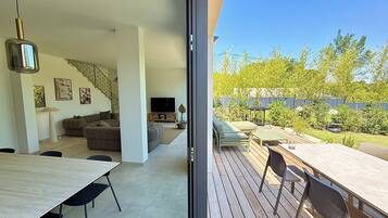 Un intérieur moderne avec un canapé, un téléviseur et un coin repas, ainsi qu’une terrasse en bois avec des meubles d’extérieur.