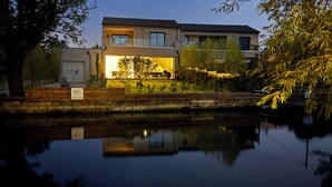 A modern two-story house with a visible garage door, a well-maintained lawn, and a calm body of water reflecting the building. - La Villa sur la Sorgue: Waterfront and Downtown in L'Isle-sur-la-Sorgue (L'Isle-sur-la-Sorgue)