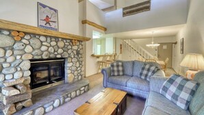 A stone fireplace, a blue sofa, and a wooden coffee table in a living room with a high ceiling and exposed beams. - Updated condo w/ shared Pools, Hot Tubs, Saunas, Fitness Room & Tennis Courts (Mammoth Lakes)