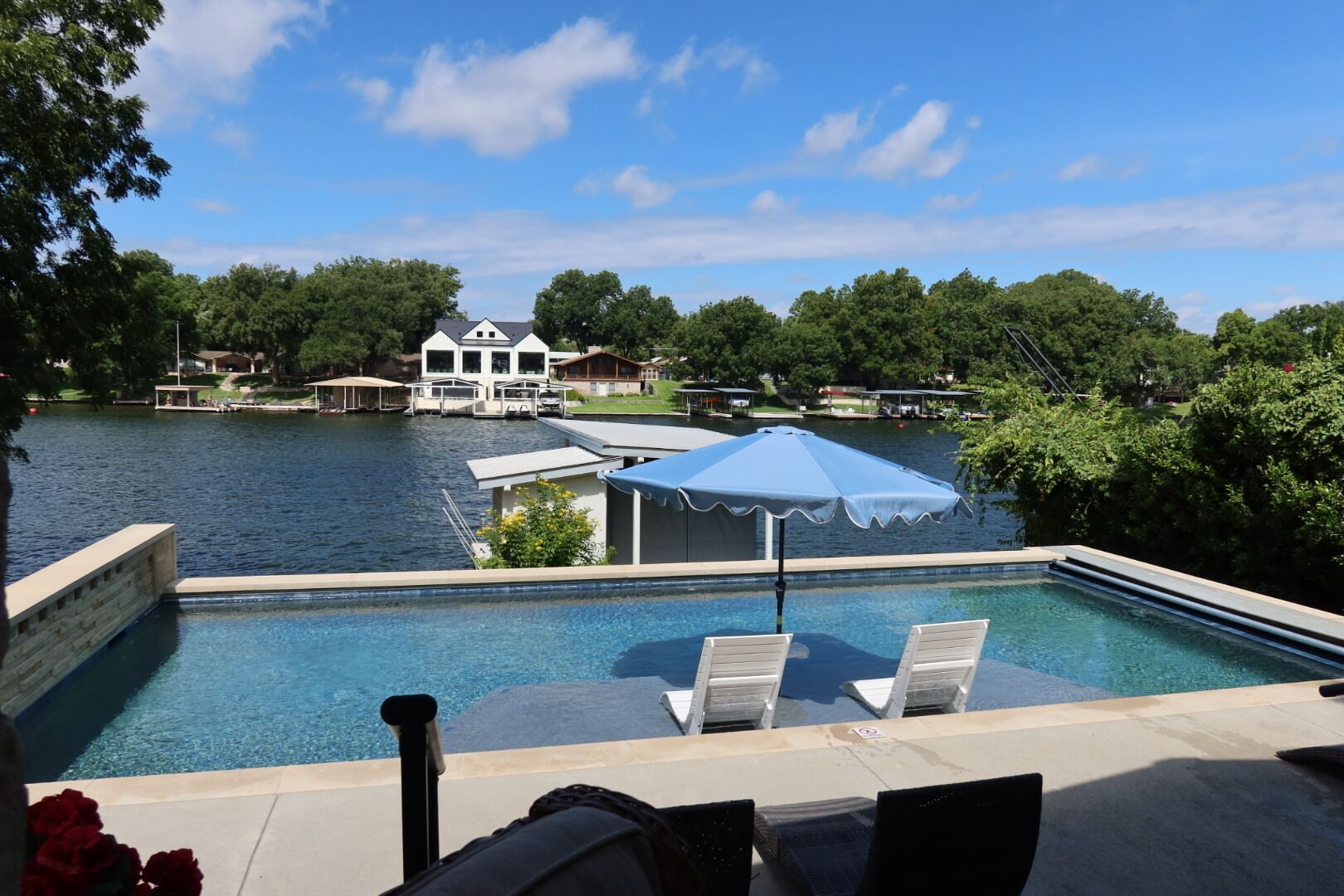 A swimming pool with lounge chairs and an umbrella, surrounded by a lake and houses in the background.