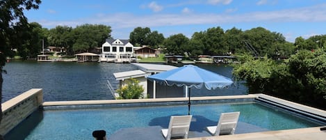 A swimming pool with lounge chairs and an umbrella, surrounded by a lake and houses in the background.