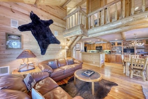 Ein rustikales Blockhaus-Interieur mit Ledersofa, rundem Couchtisch, aufgenageltem Fellmantel, Holzmöbeln und einer Küchenzeile.