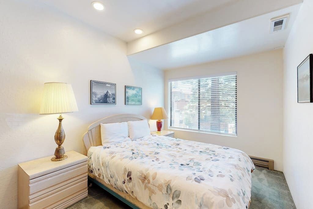 A bedroom with a bed, two bedside tables, a window with blinds, and a wall-mounted picture.