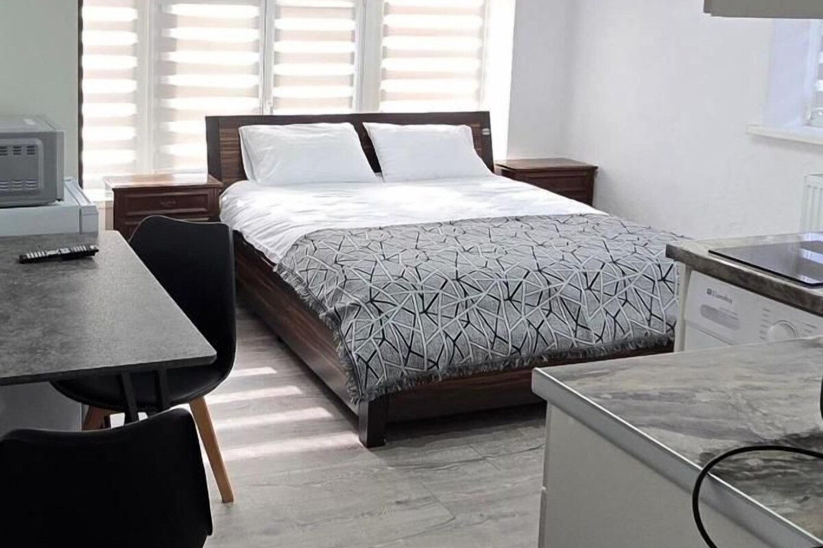 A bedroom with a bed, a desk, and a chair. There is a window with blinds, a radiator, and a small table with a lamp.