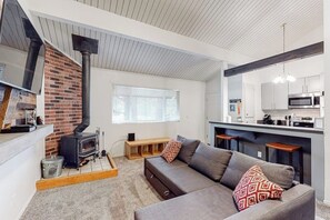 A modern living room with a fireplace, a sofa, and a kitchen area. - Charming, two-story ski condo w/ a wood stove, free WiFi, shared pool, & hot tub (Mammoth Lakes)