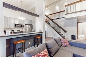 A modern kitchen with a bar area, a staircase, and a living room with a sofa and cushions. - Charming, two-story ski condo w/ a wood stove, free WiFi, shared pool, & hot tub (Mammoth Lakes)