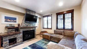 Una sala de estar con chimenea de piedra, un televisor de pantalla plana, un amplio sofá modular y una ventana con persianas.