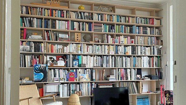 Huoneessa on suuri kirjahylly täynnä kirjoja, televisio, rottinkituoli ja puulattia.