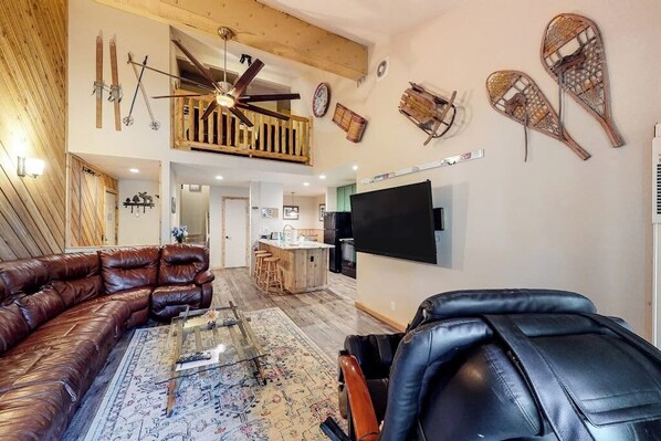 A spacious living area with a leather sofa, a black leather chair, a glass coffee table, a ceiling fan, and a mounted TV.