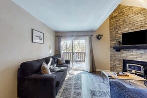 Ein Wohnzimmer mit einer Couch, einem Flachbildfernseher, einem Kamin und einem Balkon mit Blick auf Bäume.