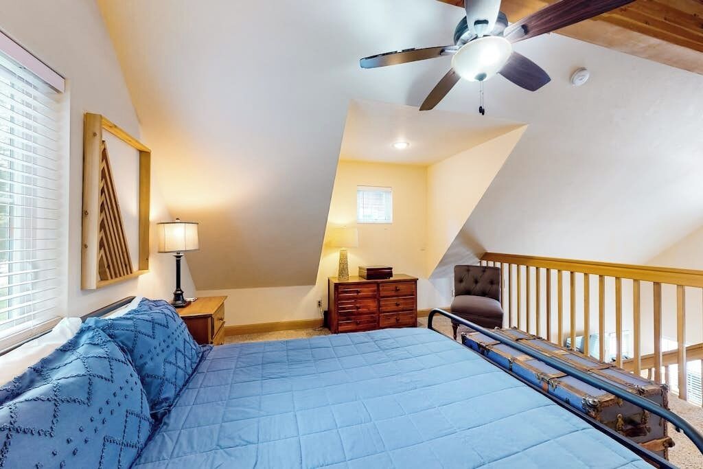 Un dormitorio con cama, ventilador de techo, mesita de noche, cómoda, silla y escalera.
