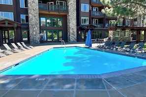 A swimming pool with lounge chairs and a stone building with balconies. - Beautiful & Convenient Suite Close to Activities w/Shared Pool & Hot Tub, WiFi (Mammoth Lakes)