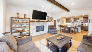 A spacious living room with a fireplace, a flat-screen TV mounted above it, and a dining area with a kitchen in the background. - Luxe, Modern Townhome w/ Mountain Views & WiFi - Close to Golf & Skiing (Mammoth Lakes)
