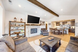 A spacious living room with a fireplace, a flat-screen TV mounted above it, and a dining area with a kitchen in the background. - Luxe, Modern Townhome w/ Mountain Views & WiFi - Close to Golf & Skiing (Mammoth Lakes)