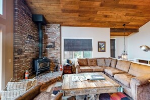 Ein gemütliches Wohnzimmer mit Backsteinwand, einem schwarzen Kaminofen, einem Holztisch und einem beigen Ecksofa.