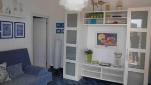 Ein Zimmer mit einem blauen Sofa, weißen Regalen mit Glastüren, einer hängenden weißen Leuchte und einer Tür zu einem weiteren Raum.
