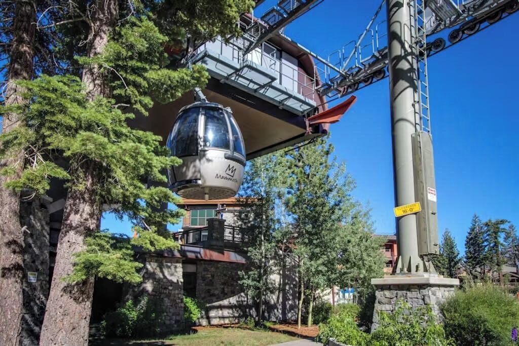 Sistema de teleférico con cabina suspendida entre dos torres, rodeado de árboles y un edificio de piedra.