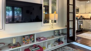 Een moderne woonkamer met een flatscreen televisie, een witte airconditioner, een raam met glas, een lamp en een boekenkast met boeken en decoratieve objecten.