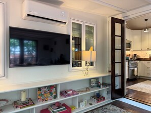 A modern living room with a flat-screen TV, a white air conditioner, a glass-fronted window, a lamp, and a bookshelf with books and decorative items. - Charming, World Cup Ready 2-bedroom Bungalow in Ashford Park, Chamblee- Near PDK (Chamblee)