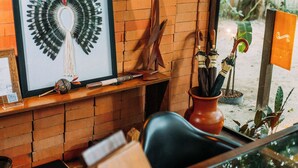 Una habitación con pared de ladrillo, una obra de arte enmarcada, estante de madera con objetos, una silla negra, una mesa con un jarrón y una ventana con vista a follaje.