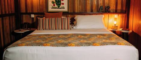 A bed with a patterned bedspread, a pillow, a bedside table, a framed cactus artwork, and a wall with wooden paneling and a letter 'A' sign.