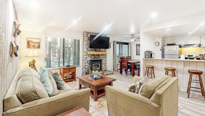 A living room with a fireplace, a dining area, and a kitchen with bar stools. - Condo Near Eagle Lodge & Golf w/ Fireplace, WiFi & Shared Pool/Hot Tub/Sauna (Mammoth Lakes)