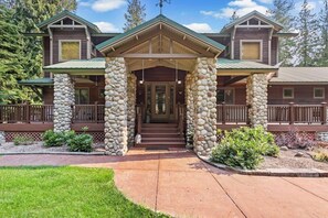 A two-story lodge with a stone entrance, wooden deck, and a clear view of the surrounding forest.
