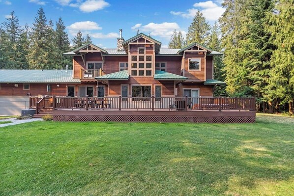 A large, two-story wooden house with a balcony, surrounded by a well-maintained lawn and tall trees.