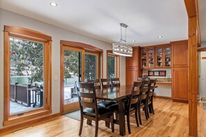 A dining area with a large wooden table and chairs, wooden cabinets with glass doors, and large windows.