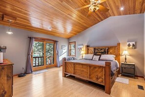 A bedroom with a wooden ceiling, a large bed, a nightstand, and a view through a sliding glass door.
